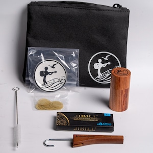 May include: A black zippered pouch with a white logo of a person playing a guitar on a surfboard. The kit includes a wooden container, a cleaning brush, a metal tool, a pack of filters, and a box of JIBILL rolling papers.