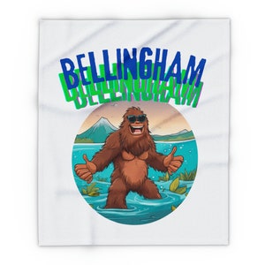 Könnte beinhalten: Eine weiße Decke mit einer Cartoon-Illustration eines lächelnden Bigfoots, der eine Sonnenbrille trägt und in einem See steht. Der Text "BELLINGHAM" ist in blauen und grünen Buchstaben über dem Bild geschrieben.