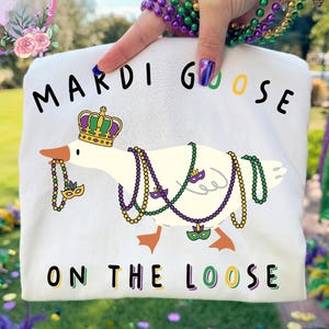 May include: White t-shirt featuring a cartoon goose adorned with a crown and Mardi Gras beads. The text "MARDI GOOSE ON THE LOOSE" is printed on the shirt. The image is held up by a person with Mardi Gras beads on their wrist.
