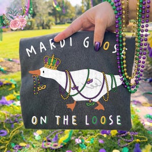 May include: A dark gray tote bag featuring a cartoon goose wearing a crown and Mardi Gras beads. The text "MARDI GOOS" and "ON THE LOOSE" are printed on the bag. The background includes green grass, beads, and decorations.