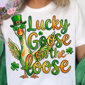 May include: White t-shirt featuring a cartoon goose wearing a green hat and shamrock glasses. The text "Lucky Goose on the Loose" is in green and orange. The design is themed for St. Patrick's Day.