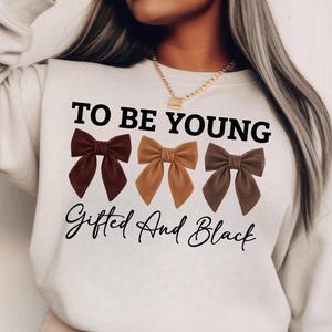 Op de afbeelding: Een cr&egrave;mekleurige sweatshirt met de tekst "TO BE YOUNG" boven drie strikjes in bruintinten. Onder de strikken staat de tekst "Gifted And Black" in een scriptlettertype. De sweatshirt heeft een casual stijl.