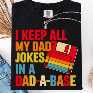 Könnte beinhalten: Schwarzes T-Shirt mit dem Text "I KEEP ALL MY DAD JOKES IN A DAD-A-BASE" in Rot, Orange, Gelb und Blau. Eine Grafik einer Diskette im Retro-Stil ist ebenfalls abgebildet. Das Shirt ist von der Marke Comfort Colors.