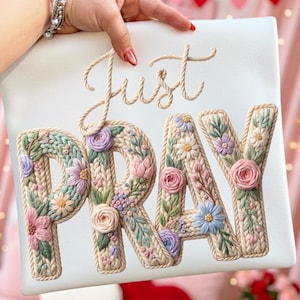 May include: A white cushion featuring the embroidered words "Just PRAY" in a beige, rope-like font. The letters of "PRAY" are adorned with colourful floral patterns in pink, purple, and blue. The cushion is held by a hand.