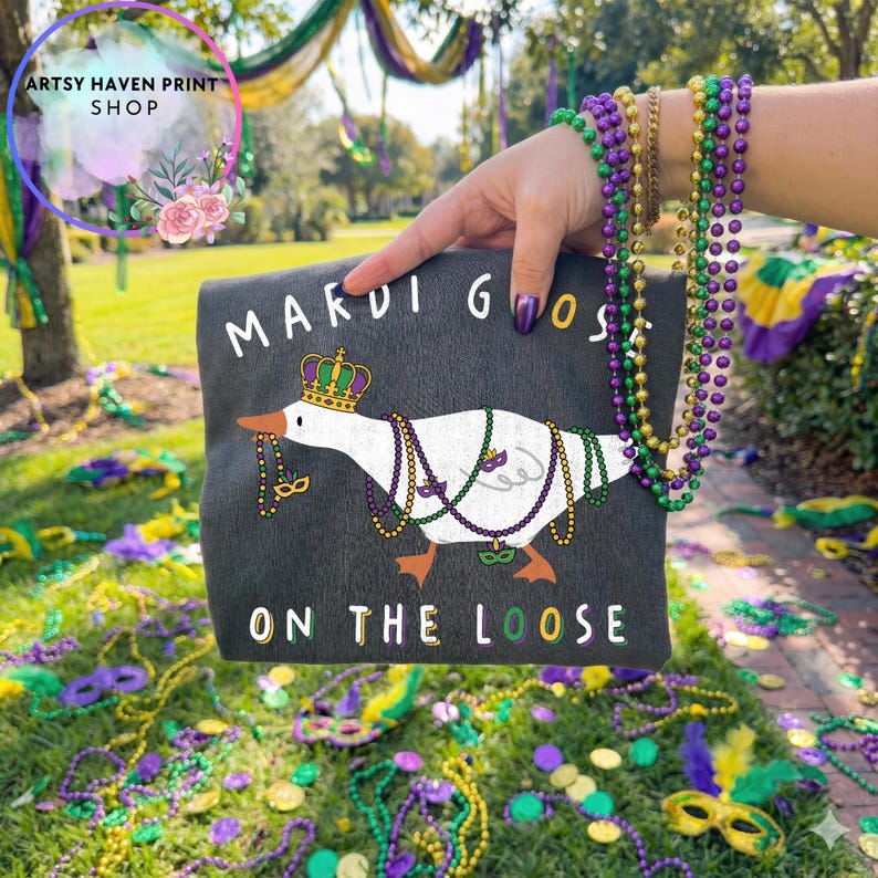 May include: A dark gray tote bag featuring a cartoon goose wearing a crown and Mardi Gras beads. The text "MARDI GOOS" and "ON THE LOOSE" are printed on the bag. The background includes green grass, beads, and decorations.
