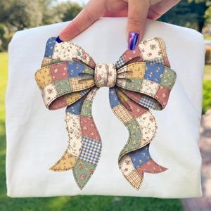 May include: A decorative bow with a patchwork design. The bow features a variety of colors, including blue, yellow, pink, and green, arranged in a quilted pattern. The bow is held up by a hand, and the background is blurred.