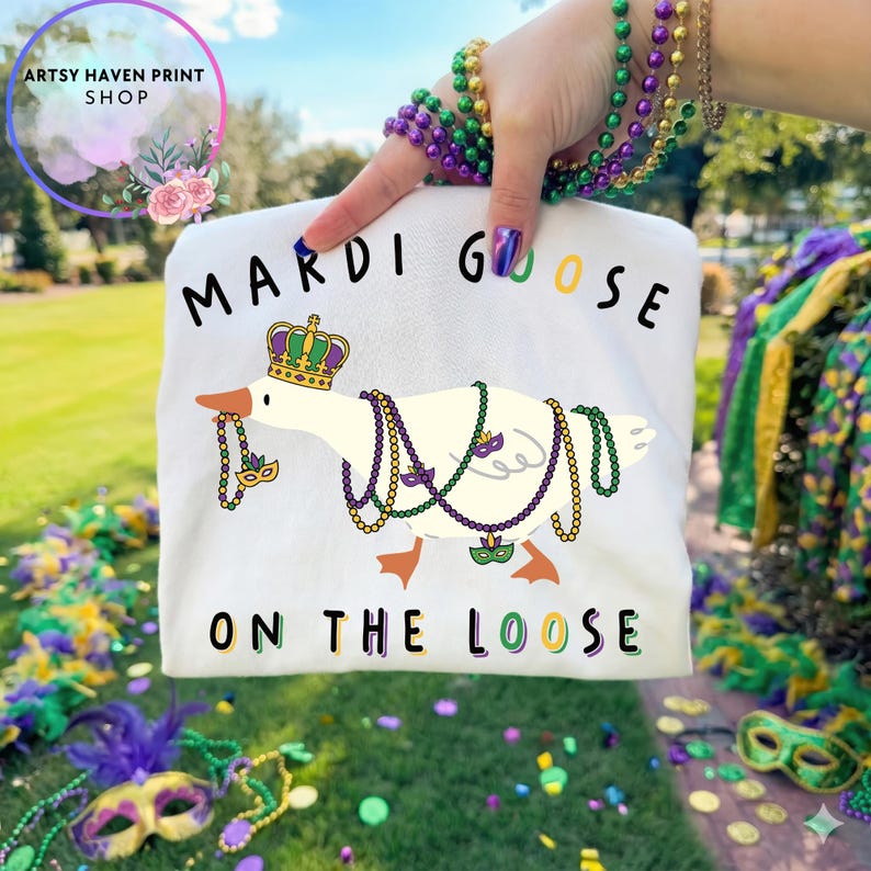 May include: White t-shirt featuring a cartoon goose adorned with a crown and Mardi Gras beads. The text "MARDI GOOSE ON THE LOOSE" is printed on the shirt. The image is held up by a person with Mardi Gras beads on their wrist.