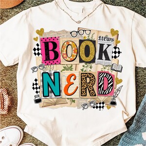 May include: Cream-coloured t-shirt featuring a vibrant "BOOK NERD" design. The letters are decorated with diverse patterns, such as polka dots, floral, and animal prints. The design incorporates glasses, hearts, and open books.