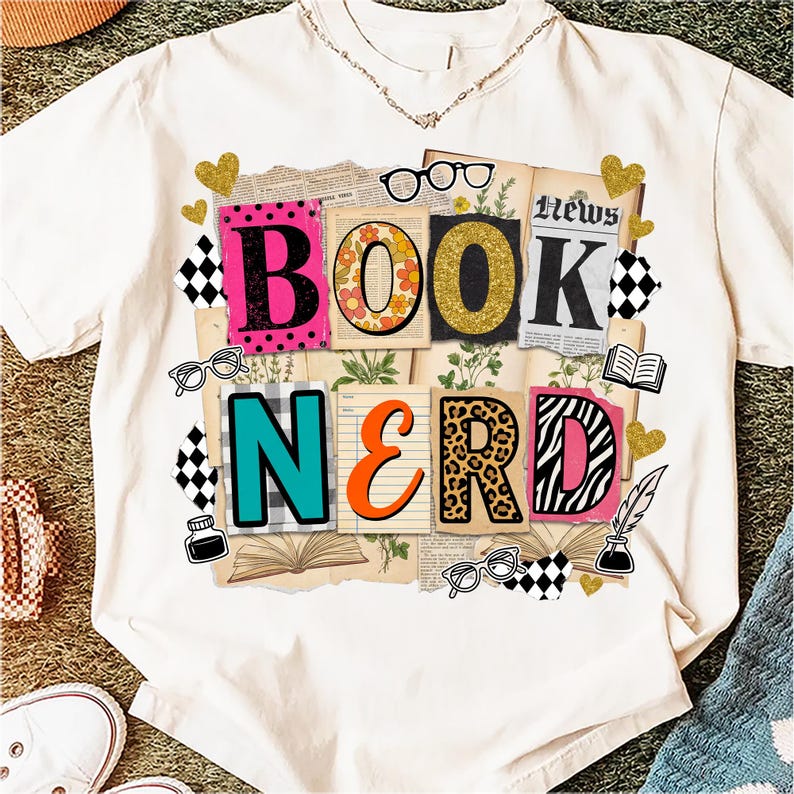 May include: Cream-coloured t-shirt featuring a vibrant "BOOK NERD" design. The letters are decorated with diverse patterns, such as polka dots, floral, and animal prints. The design incorporates glasses, hearts, and open books.
