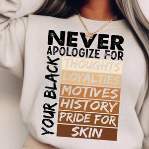 May include: A white sweatshirt with the text "NEVER APOLOGIZE FOR YOUR BLACK" in black lettering. The text is stacked vertically with the words "THOUGHTS", "LOYALTIES", "MOTIVES", "HISTORY", "PRIDE FOR", and "SKIN" in a rainbow of colours.