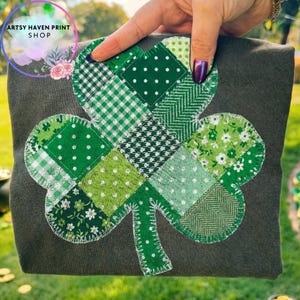 May include: A dark grey t-shirt with a large, patchwork shamrock design in various shades of green. The shamrock is made of different patterned fabrics, including polka dots, gingham, and floral prints. The text "ARTSY HAVEN PRINT SHOP" is in a circle in the upper left.