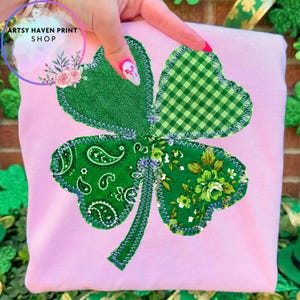 May include: A pink sweatshirt featuring a large, appliqued shamrock. The shamrock is composed of four green leaves, each with a different pattern: denim, gingham, paisley, and floral. The text "ARTSY HAVEN PRINT SHOP" is in a circle in the upper left.