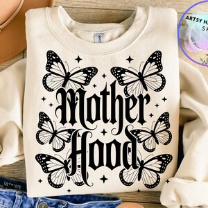 Puede incluir: Sudadera color crema con la palabra "Motherhood" en fuente g&oacute;tica, rodeada de seis ilustraciones de mariposas en blanco y negro y detalles de estrellas. La sudadera se muestra sobre una superficie plana.