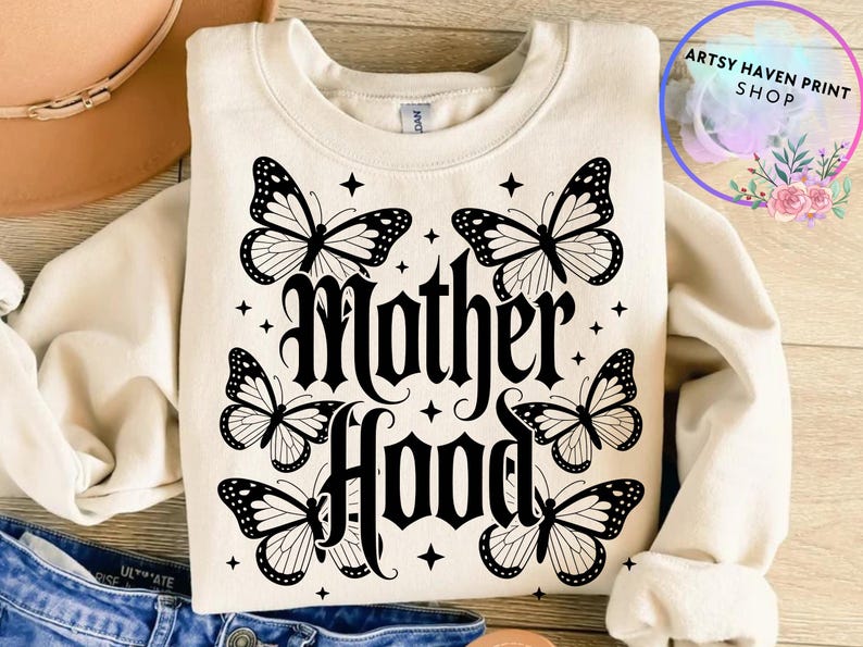 Puede incluir: Sudadera color crema con la palabra "Motherhood" en fuente g&oacute;tica, rodeada de seis ilustraciones de mariposas en blanco y negro y detalles de estrellas. La sudadera se muestra sobre una superficie plana.