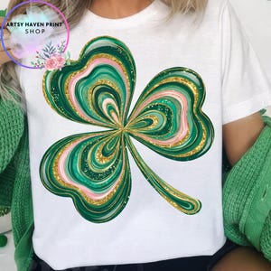 K&ouml;nnte beinhalten: Wei&szlig;es T-Shirt mit einem gro&szlig;en, farbenfrohen Kleeblatt-Design. Das Kleeblatt hat gr&uuml;ne, rosa und goldene Glitzerakzente. Das Shirt ist ein l&auml;ssiger Stil, perfekt f&uuml;r St. Patrick's Day oder den Alltag. Der Text "ARTSY HAVEN PRINT SHOP" ist sichtbar.