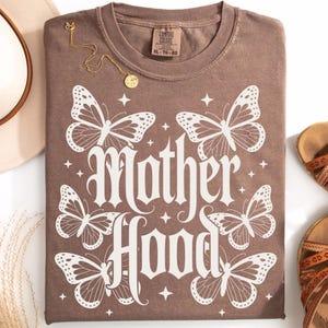 Puede incluir: Una camiseta marr&oacute;n con la palabra "Motherhood" en fuente g&oacute;tica, rodeada de ilustraciones de mariposas blancas. Un collar dorado con un colgante est&aacute; sobre la camiseta. La camiseta est&aacute; doblada y lista para usar.
