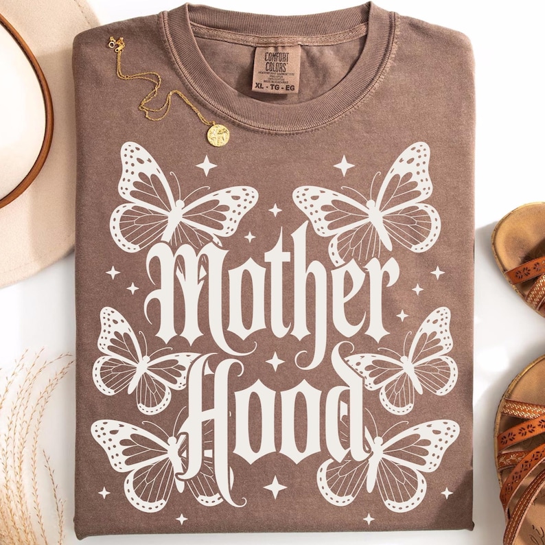 Puede incluir: Una camiseta marr&oacute;n con la palabra "Motherhood" en fuente g&oacute;tica, rodeada de ilustraciones de mariposas blancas. Un collar dorado con un colgante est&aacute; sobre la camiseta. La camiseta est&aacute; doblada y lista para usar.