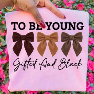 Op de afbeelding: Een lichtroze sweatshirt met de tekst "TO BE YOUNG" boven drie bruine strikken en de woorden "Gifted And Black" in cursief. De strikken zijn in bruin- en beigetinten.