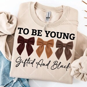 Op de afbeelding: Een beige sweatshirt met de tekst "TO BE YOUNG Gifted And Black" in het zwart. Drie bruine strik-afbeeldingen staan onder de tekst. Een zilveren ketting is zichtbaar bij de kraag.