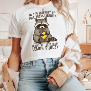 May include: White t-shirt with a graphic of a raccoon holding a lemon. The text on the shirt reads "In the interest of full transparency it was not easy peasy lemon squeezy."