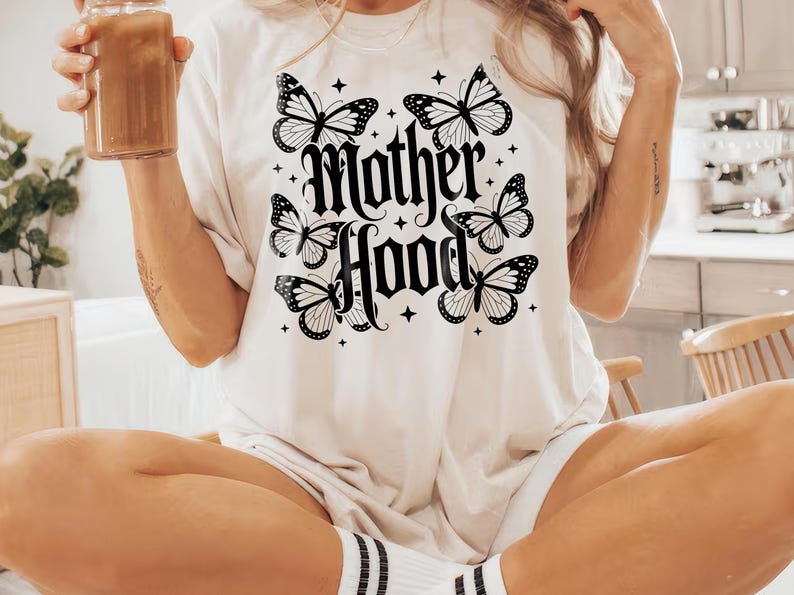 Puede incluir: Camiseta color crema con la palabra "Motherhood" en fuente g&oacute;tica, rodeada de seis mariposas en blanco y negro y peque&ntilde;os acentos de estrellas. La camiseta es de estilo casual y c&oacute;modo.