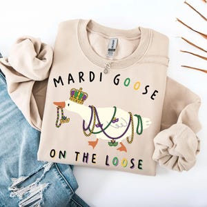 May include: Beige sweatshirt with a graphic of a goose wearing a crown and Mardi Gras beads. The text "MARDI GOOSE ON THE LOOSE" is printed above and below the goose in black, green, and purple.