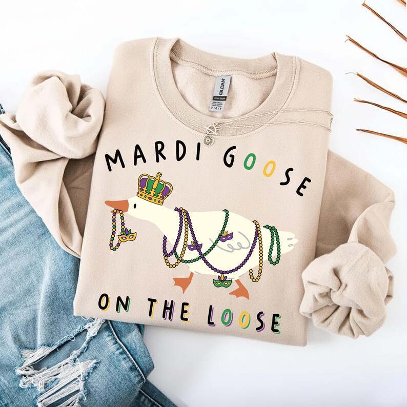 May include: Beige sweatshirt with a graphic of a goose wearing a crown and Mardi Gras beads. The text "MARDI GOOSE ON THE LOOSE" is printed above and below the goose in black, green, and purple.