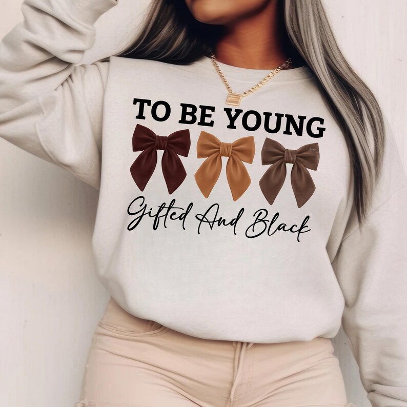 Op de afbeelding: Een cr&egrave;mekleurige sweatshirt met de tekst "TO BE YOUNG" boven drie strikjes in bruintinten. Onder de strikken staat de tekst "Gifted And Black" in een scriptlettertype. De sweatshirt heeft een casual stijl.