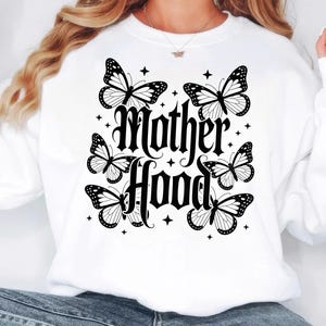 Puede incluir: Sudadera blanca con las palabras "Mother Hood" en fuente g&oacute;tica, rodeada de mariposas negras y blancas y peque&ntilde;os acentos de estrellas. Una prenda casual y c&oacute;moda.