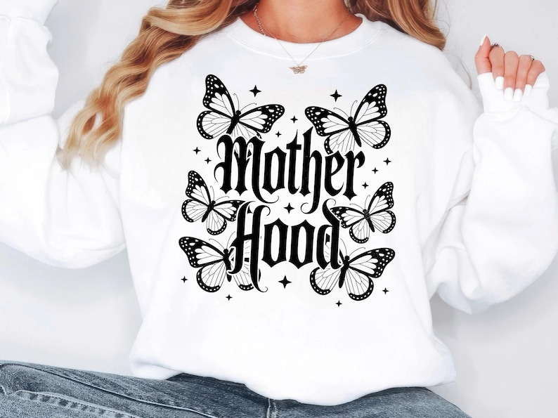 Puede incluir: Sudadera blanca con las palabras "Mother Hood" en fuente g&oacute;tica, rodeada de mariposas negras y blancas y peque&ntilde;os acentos de estrellas. Una prenda casual y c&oacute;moda.