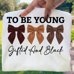 Op de afbeelding: Wit t-shirt met de tekst "TO BE YOUNG" boven drie bruine strikken. Daaronder staat in script "Gifted And Black". Het t-shirt wordt door een hand omhoog gehouden, met een groene achtergrond.
