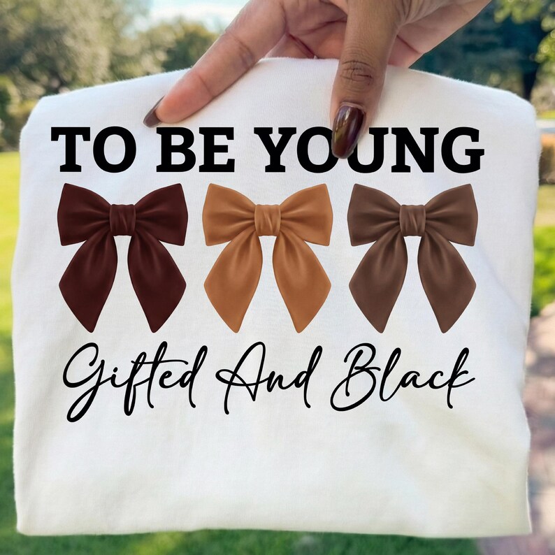 Op de afbeelding: Wit t-shirt met de tekst "TO BE YOUNG" boven drie bruine strikken. Daaronder staat in script "Gifted And Black". Het t-shirt wordt door een hand omhoog gehouden, met een groene achtergrond.