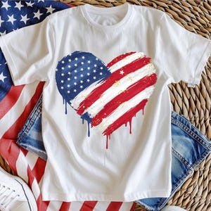 May include: White t-shirt featuring a red, white and blue heart-shaped American flag design. The flag is painted in a dripping style with gold glitter accents.