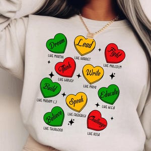 Può includere: Una felpa bianca con un design grafico di cuori che presentano parole come "Dream", "Lead", "Fight", "Think", "Write", "Build", "Believe", "Speak", "Educate" e "Challenge". Ogni cuore è di un colore diverso: rosso, giallo, verde o una combinazione di questi colori. Le parole "Like Martin", "Like Harriet", "Like Malcolm", "Like Garvey", "Like Maya", "Like Madam CJ", "Like Thurgood", "Like Frederick", "Like WEB" e "Like Rosa" sono scritte sotto ogni cuore.