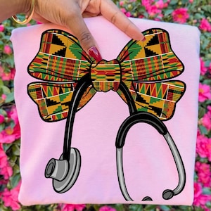 Può includere: Una felpa rosa chiaro con un design a fiocco colorato con un motivo Kente e una grafica di stetoscopio nero. L'arco è nei toni del rosso, verde, giallo e nero.