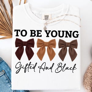 Op de afbeelding: Wit T-shirt met de tekst "TO BE YOUNG" boven drie bruine strikken en de tekst "Gifted And Black". Een zilveren ketting met een bloemhanger ligt op het shirt.
