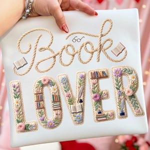 May include: A white rectangular pillow with the words "Book Lover" embroidered in a rope-like design. The letters are decorated with flowers, books, and a quill. The word "Book" has a pair of glasses above it.