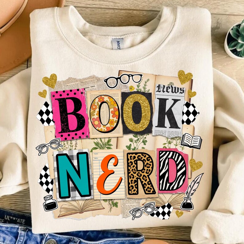 May include: Cream sweatshirt with a colourful "Book Nerd" design. The design features the words "BOOK" and "NERD" in various fonts and patterns, including floral, leopard print, and zebra stripes, with glasses and hearts.