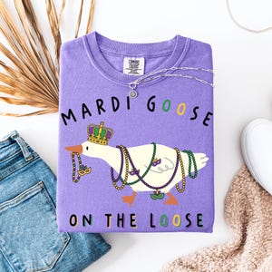 May include: A purple sweatshirt with the words "MARDI GOOSE ON THE LOOSE" and a cartoon goose wearing a crown and Mardi Gras beads. The sweatshirt is next to a pair of blue jeans and a white shoe.