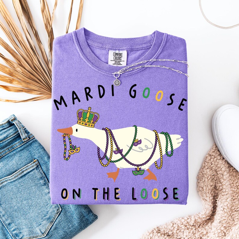May include: A purple sweatshirt with the words "MARDI GOOSE ON THE LOOSE" and a cartoon goose wearing a crown and Mardi Gras beads. The sweatshirt is next to a pair of blue jeans and a white shoe.