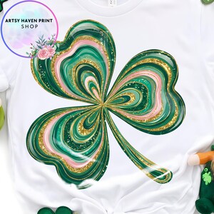 K&ouml;nnte beinhalten: Wei&szlig;es T-Shirt mit einem gro&szlig;en, stilisierten Kleeblatt-Design. Das Kleeblatt hat gr&uuml;ne, rosa und goldene Glitzerakzente. Das Shirt hat den Text "Artsy Haven Print Shop" in einem kreisf&ouml;rmigen Design mit Blumen.