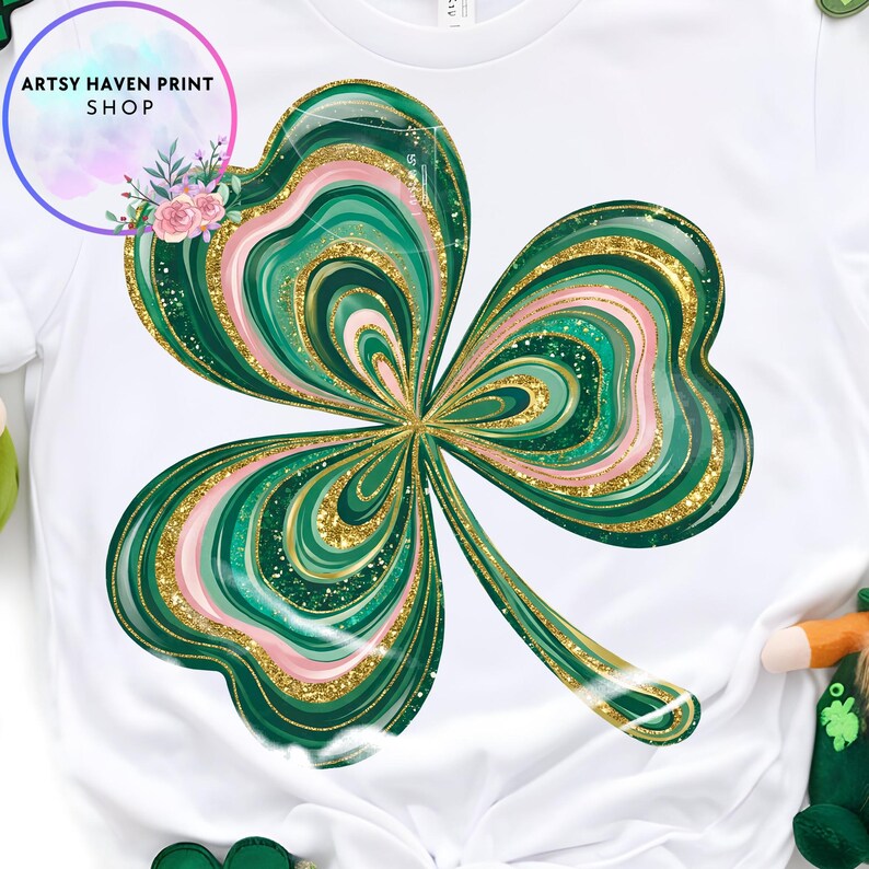 K&ouml;nnte beinhalten: Wei&szlig;es T-Shirt mit einem gro&szlig;en, stilisierten Kleeblatt-Design. Das Kleeblatt hat gr&uuml;ne, rosa und goldene Glitzerakzente. Das Shirt hat den Text "Artsy Haven Print Shop" in einem kreisf&ouml;rmigen Design mit Blumen.