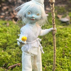 May include: A whimsical doll with pale blue skin, white hair, and a serene expression. It wears a white outfit and holds a staff and a small yellow object. The doll is set against a backdrop of green moss and foliage.