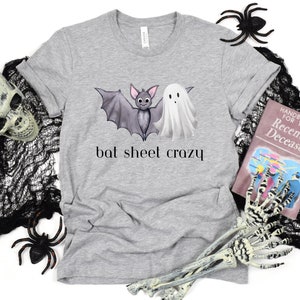 Peut inclure: Un t-shirt gris avec un motif de chauve-souris et de fantôme et le texte "bat sheet crazy". Le t-shirt est entouré de décorations d'Halloween, notamment un crâne, des toiles d'araignées et un livre intitulé "Handbook for the Recently Deceased".