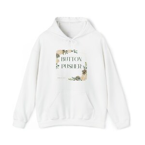 Puede incluir: Sudadera con capucha blanca con un diseño floral y el texto "BUTTON PUSHER" en un marco dorado.