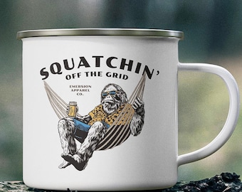 Puede incluir: Taza de camping de esmalte blanco con un gráfico de un Sasquatch de dibujos animados con gafas de sol y relajándose en una hamaca mientras bebe una cerveza. El texto en la taza dice "Squatchin' Off The Grid" y "Emerson Apparel Co."