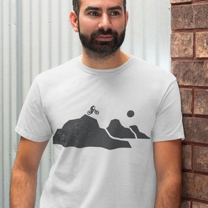 K&ouml;nnte beinhalten: Ein wei&szlig;es T-Shirt mit einer schwarzen Silhouette einer Bergkette und einer Person, die mit dem Fahrrad &uuml;ber die Berge f&auml;hrt. Die Sonne ist ebenfalls in der Silhouette zu sehen.