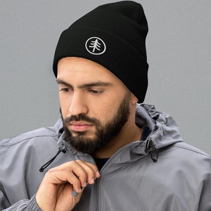 May include: A black beanie with a white embroidered tree logo. The beanie is worn by a person with a beard and a gray jacket.