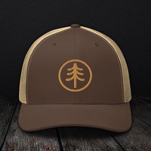 Può includere: Un cappello da camionista marrone e beige con un logo di albero ricamato marrone in un cerchio. Il cappello ha una rete sul retro.