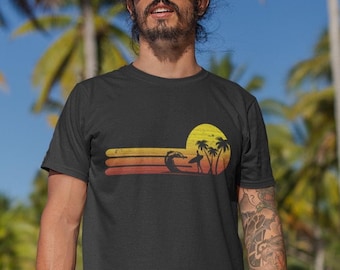 May include: Black t-shirt with a retro sunset graphic featuring a surfer, palm trees, and a sun. The graphic is in shades of orange, yellow, and red.